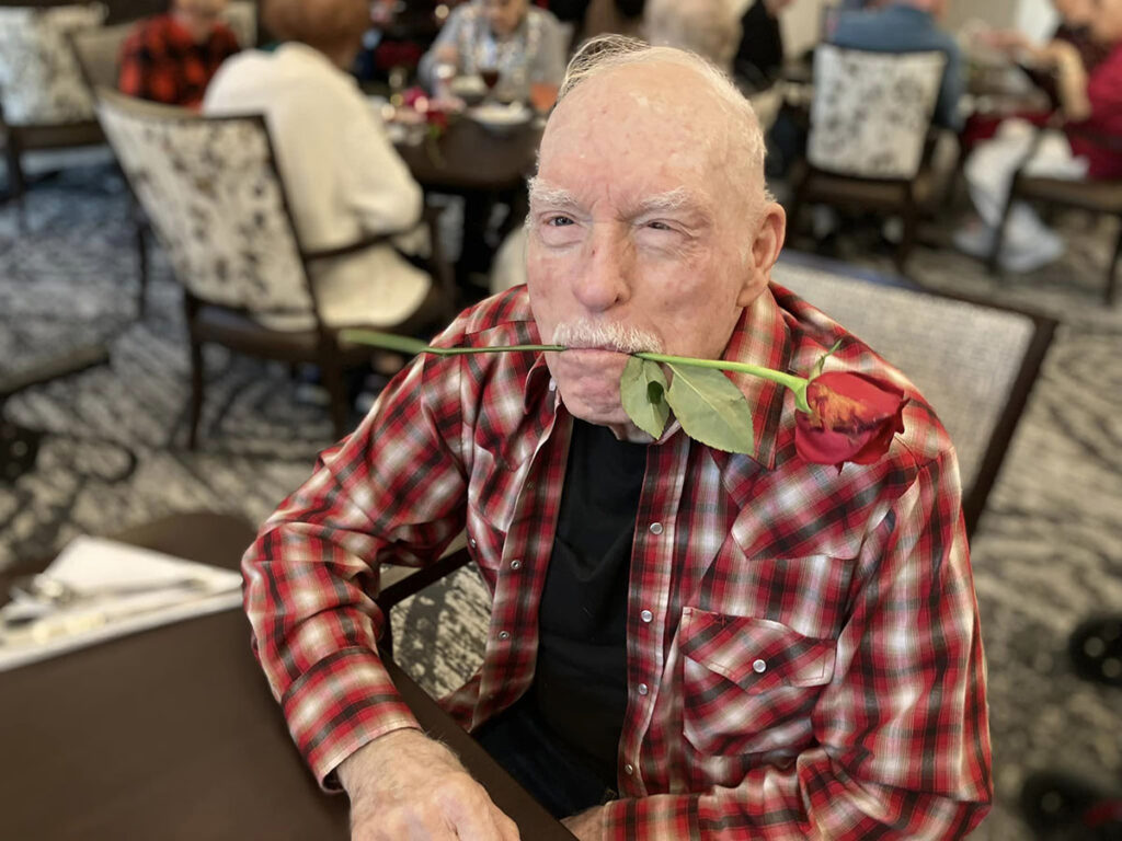Senior resident enjoying a Candle Light Dinner, playfully holding a red rose in his mouth with a joyful expression.