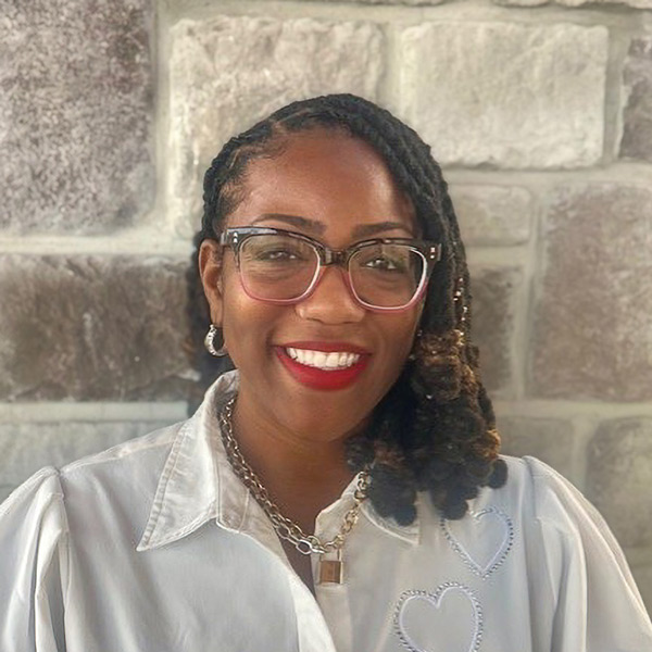 Jasmine Davis, Operations Coordinator at The Boulevard Senior Living of Saint Peters, smiling in a professional headshot. She has braided hair, wears large glasses, red lipstick, and a white blouse with heart stitching, standing in front of a light stone wall background.