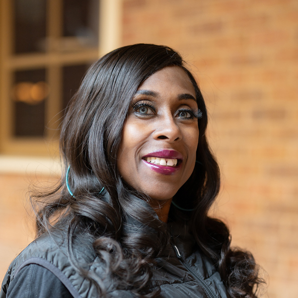 Carla Dennis, Culinary Director at The Boulevard Senior Living of Saint Peters, smiling in a professional headshot, wearing a black vest and hoop earrings, with a softly blurred brick background.