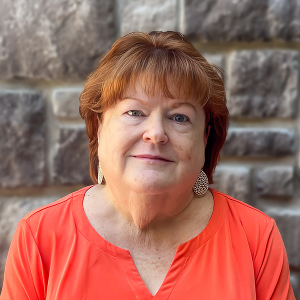 Renee Shea, Senior Living Coordinator at The Boulevard Senior Living of St. Peters, looking at the camera in a professional headshot with short auburn hair, drop earrings, a coral blouse, and a gray stone wall background.