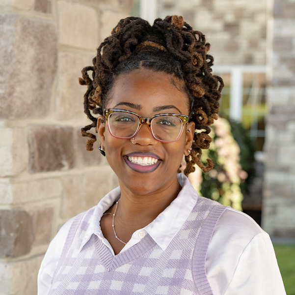 Jasmine “Jas” Davis, Operations Coordinator at The Boulevard Senior Living of St. Peters, smiling in a professional outdoor headshot with locs styled up, patterned glasses, a lavender sweater vest, and a softly blurred stone building background.