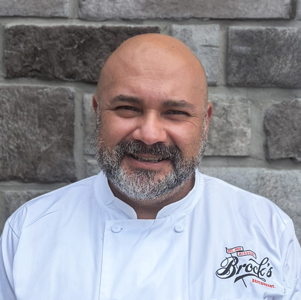 Oscar Aceituno, Culinary Director at The Boulevard Senior Living of St. Peters, smiling in a professional headshot wearing a white chef coat against a gray stone wall background.